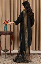 A woman wearing a black traditional outfit with a sheer black dupatta featuring gold embroidery, standing in front of beige curtains next to a small wooden table with a vase of flowers.