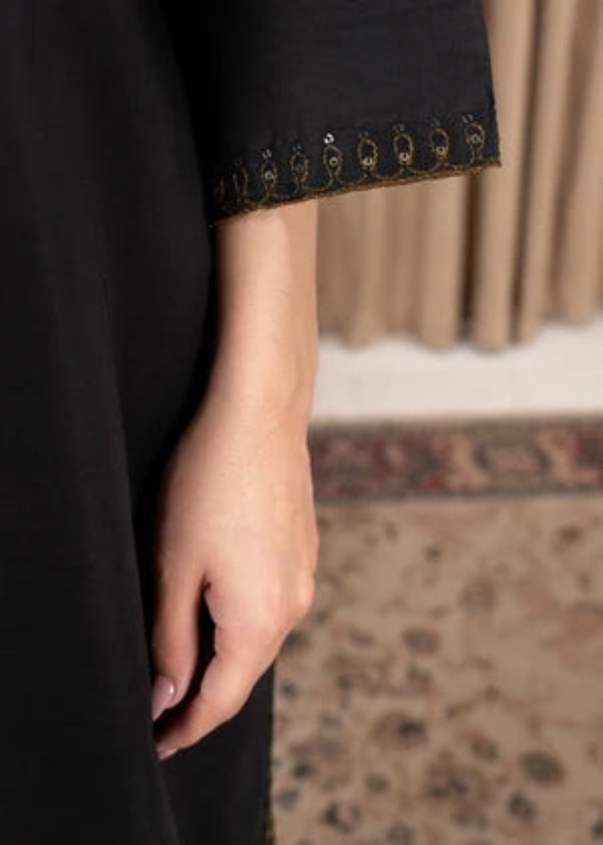 Close-up of a person wearing a black abaya with gold embroidered detailing along the sleeve cuff, with a hand resting near the fabric.